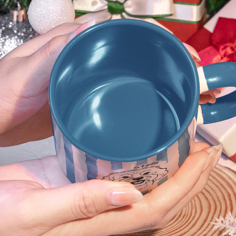 May include: Blue and white striped ceramic mug with a dog illustration and the name "Stella". The mug has a blue interior and handle. The mug is held in a person's hands.