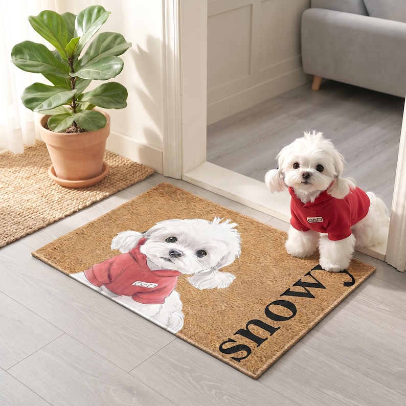 Puede incluir: Un felpudo rectangular de fibra de coco color canela con una ilustraci&oacute;n impresa de un perro blanco con una sudadera roja con capucha. El perro tiene una peque&ntilde;a etiqueta blanca en el pecho. La palabra "snowy" est&aacute; impresa en letras negras grandes.