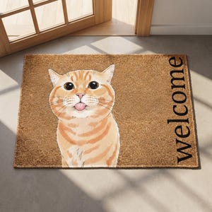 Puede incluir: Un felpudo rectangular de fibra de coco marr&oacute;n con una ilustraci&oacute;n gr&aacute;fica de un gato atigrado naranja con ojos grandes y la lengua fuera. La palabra Welcome est&aacute; impresa en letras may&uacute;sculas negras en el lado derecho de la alfombra.