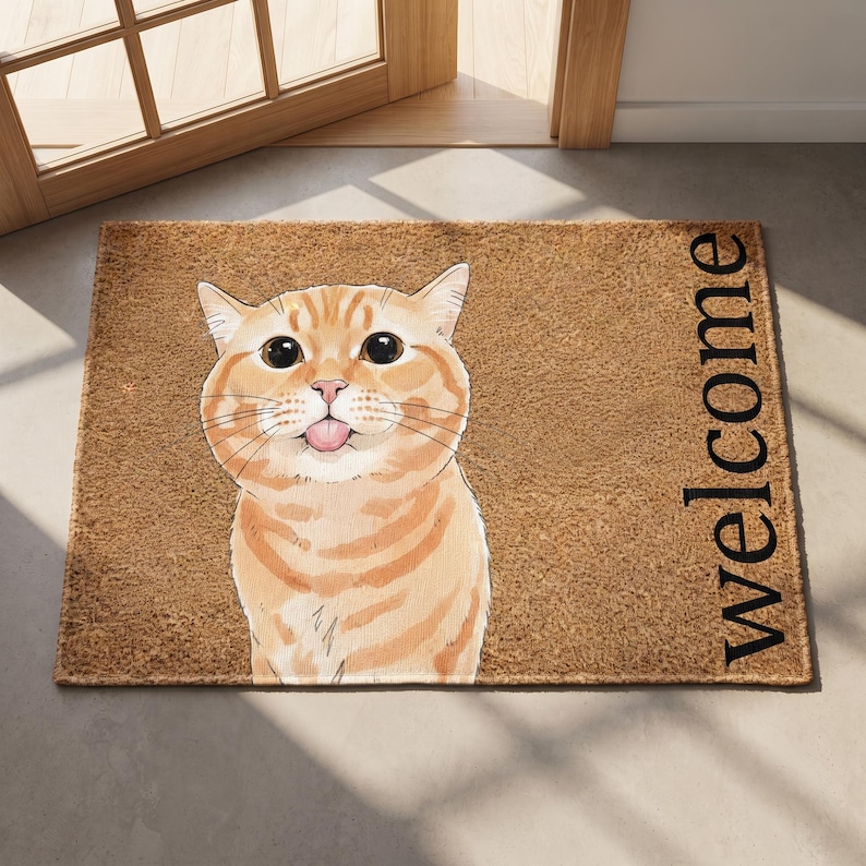 Puede incluir: Un felpudo rectangular de fibra de coco marr&oacute;n con una ilustraci&oacute;n gr&aacute;fica de un gato atigrado naranja con ojos grandes y la lengua fuera. La palabra Welcome est&aacute; impresa en letras may&uacute;sculas negras en el lado derecho de la alfombra.