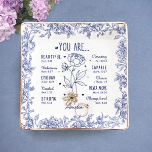 May include: A square, white ceramic trinket dish with a blue floral design and gold trim. The dish features the words "YOU ARE..." and inspirational words. Two rings and a flower-shaped jewelry piece rest on the dish.