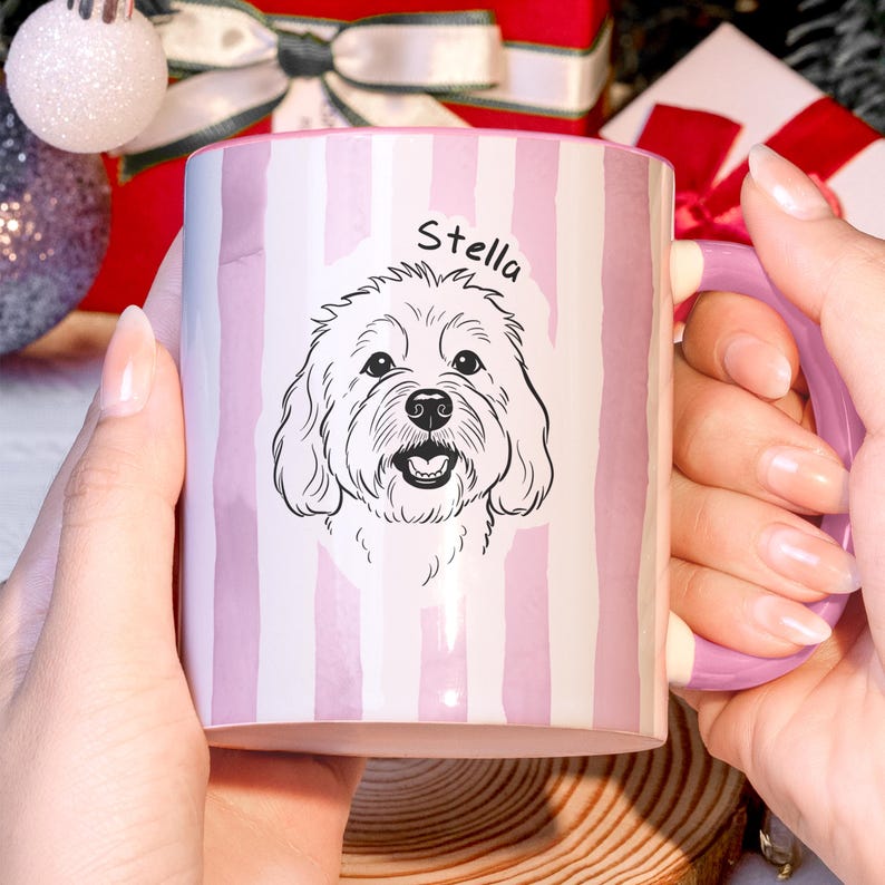 May include: A ceramic mug with a pink and white striped pattern. The mug features a black and white illustration of a dog with the name "Stella" above it. The mug has a pink handle and is held by a person.