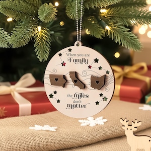 以下が含まれることがあります： 「When you are Family the miles don't matter」のテキストが入った丸い木製のクリスマスオーナメント。ハート型の切り抜きがある3つの州の切り抜きが特徴です。銀色のチェーンから吊り下げられています。
