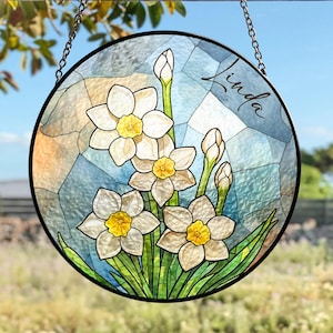 Peut inclure: Vitrail rond suspendu avec un motif floral de jonquilles blanches aux centres jaunes et des feuilles vertes. Le nom "Linda" est écrit en cursive dans le coin supérieur droit.