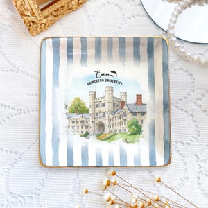 May include: Square ceramic trinket dish with blue and white vertical stripes and gold trim. The center features a watercolor illustration of Princeton University with the name "Emma" above the university name.