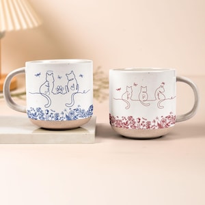 May include: Two ceramic mugs featuring cat illustrations. One mug has blue floral and cat designs with the names Simba and Cleo. The other mug has red floral and cat designs with the names Simba, Cleo, and Ingrid.