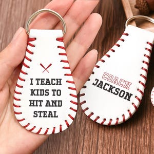 May include: White leather baseball-shaped keyrings with red stitching and silver key rings. One keyring reads "I TEACH KIDS TO HIT AND STEAL" and the other says "COACH JACKSON".