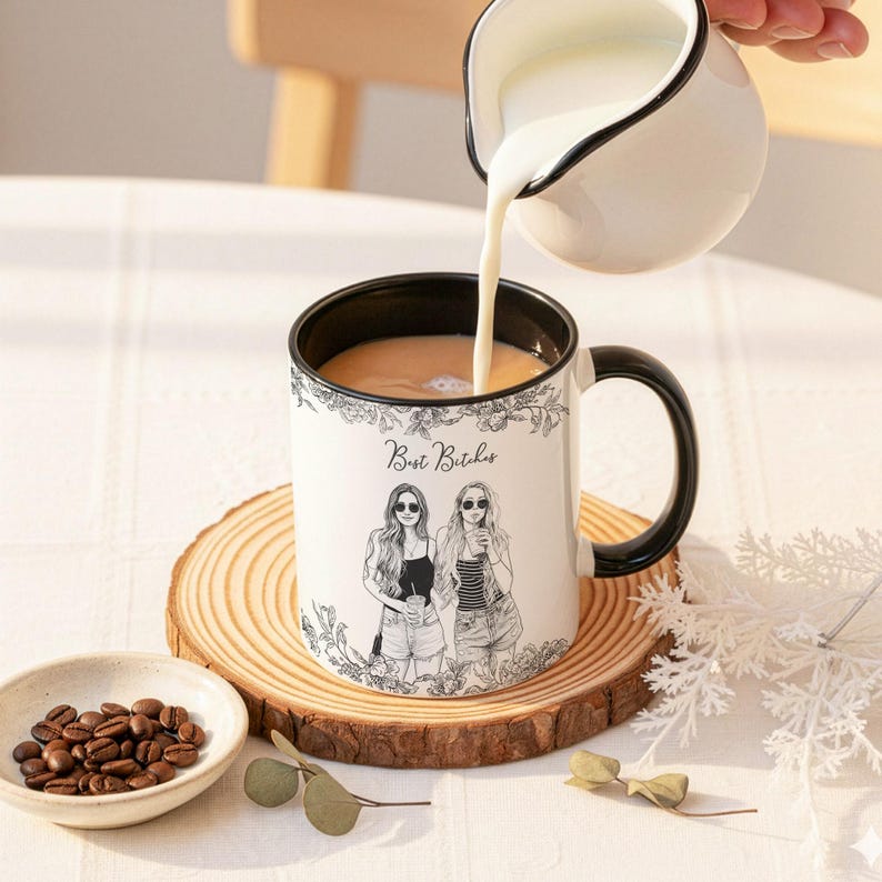 May include: A white ceramic mug with a black interior and handle, featuring a floral design and the text "Best Bitches." The mug is on a wooden coaster, with coffee beans in a small bowl and milk being poured from a white pitcher.