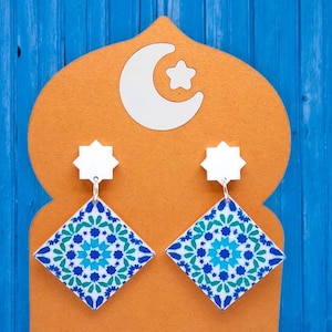 Puede incluir: Un par de pendientes con estampado azul y blanco con un diseño de estrella blanca. Los pendientes se muestran sobre un fondo naranja con una luna creciente blanca.