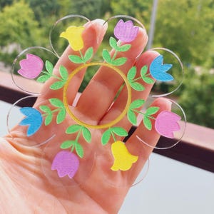 Puede incluir: Un posavasos acrílico transparente en forma de flor con un diseño floral colorido. El diseño presenta tulipanes rosas, azules y amarillos con hojas verdes y un anillo central dorado. El posavasos se sostiene en una mano.