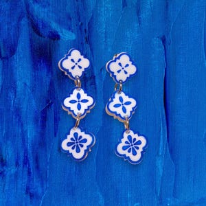 Puede incluir: Un par de pendientes colgantes con un diseño floral en blanco y azul. Cada pendiente tiene tres formas florales conectadas con conectores de metal dorado. Los pendientes están sobre un fondo azul texturizado.