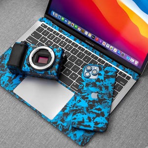 Puede incluir: Una cámara Sony, un iPhone y un ordenador portátil MacBook Air con un patrón de camuflaje azul y negro a juego. La cámara está en el panel táctil del portátil, y el teléfono está al lado del teclado. La pantalla del portátil muestra una imagen colorida.