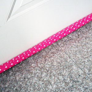 Puede incluir: Un tope de puerta de tela rosa con lunares blancos, colocado en la base de una puerta blanca. El tope está diseñado para bloquear corrientes de aire y ahorrar energía. El suelo está embaldosado con granito gris.