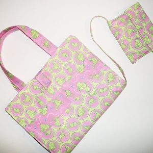 Könnte beinhalten: Eine rosa Tragetasche und eine passende Geldbörse mit einem sich wiederholenden Muster aus grünen Fröschen in rosa Kreisen. Die Tasche hat eine rechteckige Form mit Griff, und die Geldbörse hat einen langen Riemen.