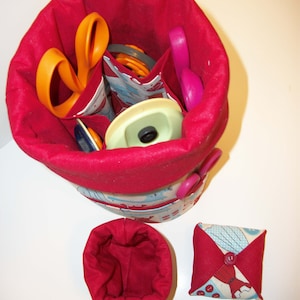 May include: A red fabric sewing basket with a variety of sewing tools inside, including orange and pink scissors. A small red fabric container and a red and patterned pin cushion are also present.