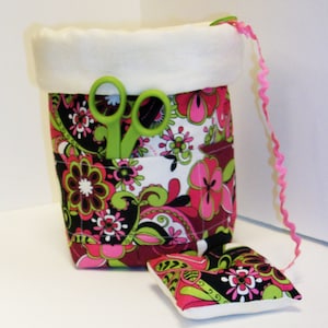 May include: A fabric sewing organizer with a floral pattern in pink, green, black, and white. The organizer has a white fleece top and a pocket holding green scissors. A matching pin cushion sits beside it, and a pink ribbon is attached.