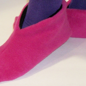 Op de afbeelding: Een paar felroze fleece pantoffels. De pantoffels hebben een levendige fuchsia kleur en een zachte, pluche textuur. De opening van de pantoffel is gevoerd met zwarte stof. Paarse sokken zijn zichtbaar in de pantoffels.