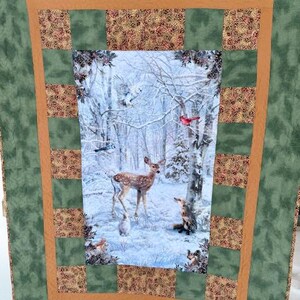 May include: A quilted blanket with a winter forest scene featuring a deer, fox, and birds. The central image is framed by a brown border and patterned squares, set against a green background.