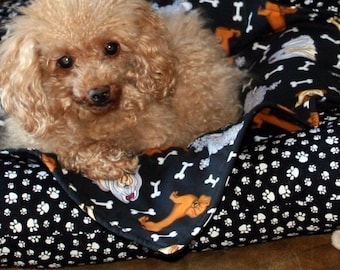 May include: A small, light brown dog rests in a black dog bed. The bed has a pattern of white paw prints and bones on a black background. The dog bed also features a pattern of orange dogs.