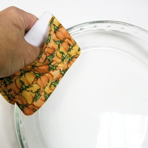 May include: A quilted pot holder with a pumpkin pattern in orange, yellow, and green is held near a clear glass pie dish. The pot holder has a white interior and is designed for kitchen use.