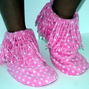 May include: Pink fleece slipper boots with white polka dots and fringe detail. These cozy boots are designed to keep feet warm and comfortable. The boots are a fun and playful accessory for lounging at home.