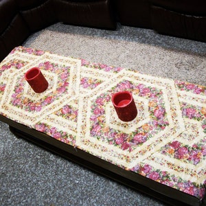 Könnte beinhalten: Ein rechteckiger Couchtisch mit floralem Muster und zwei roten Kerzenhaltern. Das florale Muster zeigt rosa und lila Blumen auf cremefarbenem Hintergrund. Der Tisch ist aus Holz.