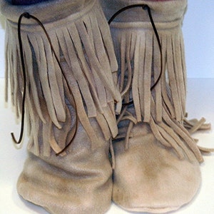 Pode incluir: Um par de botas mocassim de camurça bege com longas franjas. As botas têm uma biqueira macia e arredondada e são amarradas com um cordão castanho escuro. As franjas são de um tom bege mais claro, criando um efeito em camadas.