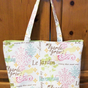 May include: White tote bag with a floral and script design. The bag features a pattern of pink, blue, and yellow flowers, birds, and the words "Le Jardin" and "my garde grows". The handles are white with a matching floral pattern.