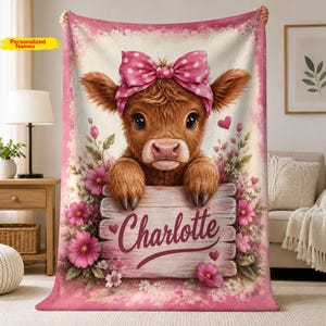 May include: A soft fleece blanket featuring a brown Highland calf with a pink polka-dot bow on its head. The calf rests its hooves on a wooden sign with the name Charlotte printed in script. The design includes pink flowers and small hearts.