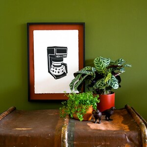 May include: A framed black and white linocut print featuring a coffee maker with a star pattern on the carafe. The artwork is displayed in a black frame with a rust-colored mat, positioned above a brown vintage trunk with metal hardware.