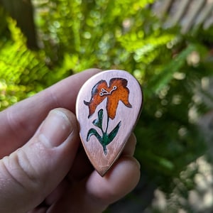 Peut inclure: Un pendentif en cuivre avec un motif de fleur orange peint à la main. La fleur a des feuilles vertes et est placée sur un fond en cuivre.