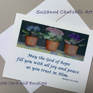 May include: A white note card with a watercolor painting of three potted African violets with blue, pink, and purple flowers. The card includes a Bible verse and comes with a white envelope. The artist's name is Suzanne Churchill.