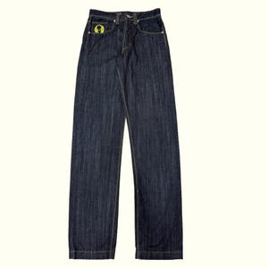 Op de afbeelding: Donkerblauwe spijkerbroek van indigo denim met een hoge taille en rechte pijpen. De broek is voorzien van een knoopsluiting, contrasterende stiksels en een kleine ronde gele patch met een zwart grafisch ontwerp op de voorzak.
