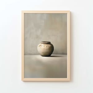 Puede incluir: Una impresión enmarcada de una naturaleza muerta con una vasija de cerámica beige y redonda con un borde oscuro. La vasija está centrada contra un fondo neutro, creando una estética minimalista. La obra de arte está dentro de un marco de madera clara.
