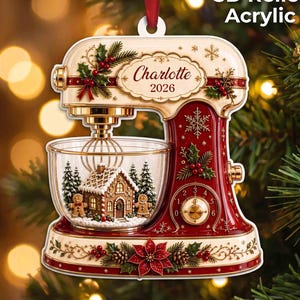 May include: A 3D relief acrylic Christmas ornament shaped like a stand mixer in cream and red. The mixer features a clear bowl containing a gingerbread house and two gingerbread people. It is decorated with holly, poinsettias, and snowflakes.