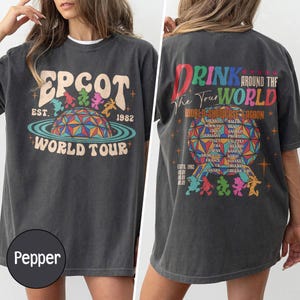 May include: A charcoal grey t-shirt featuring a front graphic with the text EPCOT EST. 1982 WORLD TOUR and a colourful dome illustration. The back graphic displays DRINK AROUND THE WORLD, WORLD SHOWCASE LAGOON, and a list of countries with cheers.