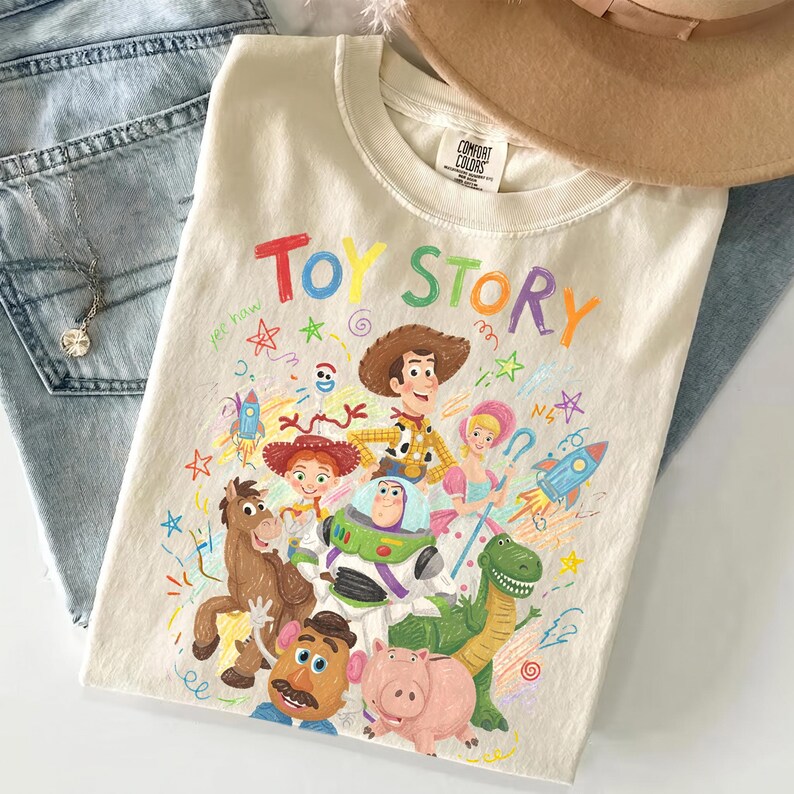 May include: A cream-colored t-shirt featuring the text "TOY STORY" in colorful letters above a graphic illustration of characters including Woody, Buzz Lightyear, Jessie, Bullseye, Rex, Hamm, Bo Peep, and Forky, surrounded by colorful doodles.