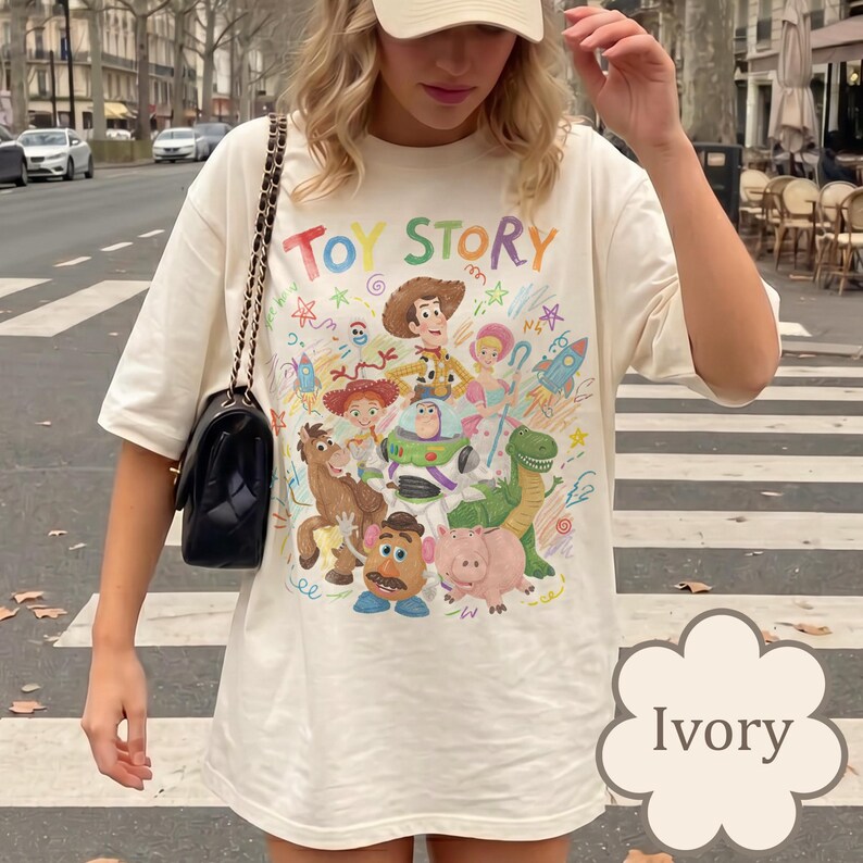 May include: An ivory t-shirt featuring a colorful graphic print of Toy Story characters including Woody, Buzz Lightyear, Jessie, Rex, Mr. Potato Head, Hamm, and Bo Peep. The text TOY STORY is printed in multicolored letters above the character group.