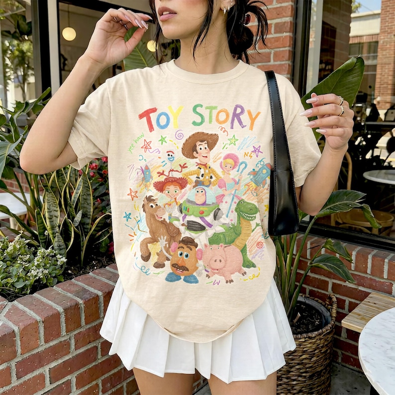 May include: A beige short-sleeve t-shirt featuring a colorful graphic print of Toy Story characters including Woody, Buzz Lightyear, Jessie, Bullseye, Mr. Potato Head, and Hamm, with the text Toy Story in rainbow-colored letters at the top.