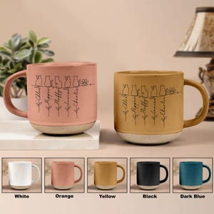 May include: Two ceramic mugs with a cat design and names. One is pink, the other is yellow, both with a white base. The mugs have a handle and are decorated with a line drawing of cats and names. Below are color options: white, orange, yellow, black, and dark blue.