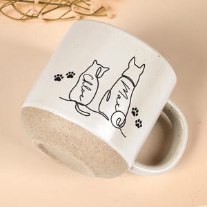 May include: Cream-colored ceramic mug with a light brown base. The mug features black line art of a cat and a dog, with the names "Chloe" and "Max" written on them. Black paw prints are scattered around the design.