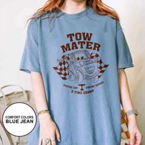 May include: A light blue t-shirt featuring a brown graphic of Tow Mater from the movie Cars, with the text "TOW MATER" above the image. Below the image are the words "Piston Cup Racing Series 5 Time Champ".