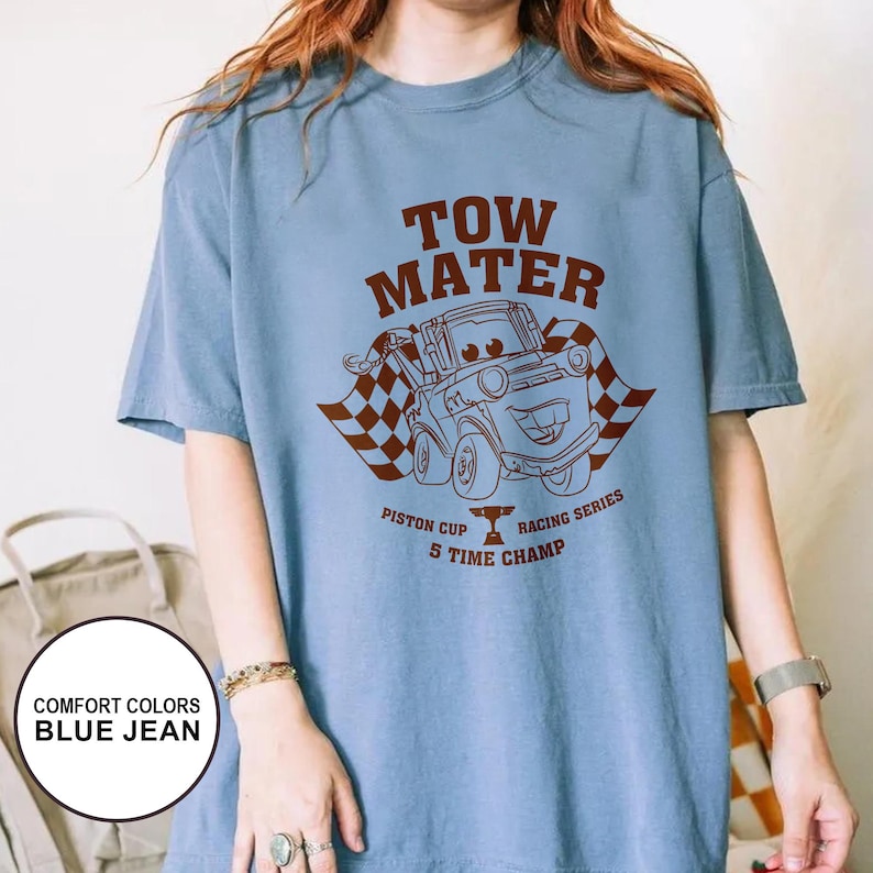 May include: A light blue t-shirt featuring a brown graphic of Tow Mater from the movie Cars, with the text "TOW MATER" above the image. Below the image are the words "Piston Cup Racing Series 5 Time Champ".