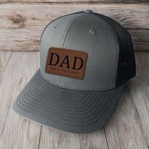 Peut inclure: Casquette de camionneur grise avec un dos en filet noir et une visière incurvée. Un patch en cuir marron sur le devant affiche le mot "DAD" en noir gras, avec "ANNA & JOHN & HENRY" en dessous. Le chapeau est posé sur un fond en bois.