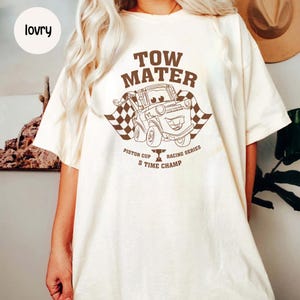 May include: Cream-colored t-shirt featuring a brown graphic of Tow Mater from the movie Cars. The graphic includes the text "TOW MATER", a checkered flag, and "PISTON CUP S TIME CHAMP".