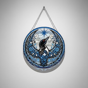 May include: Round stained glass art piece featuring a black bird with red eyes and white accents, surrounded by blue floral and leaf designs. The bird is set against a clear background with a blue starburst design. A silver chain is used for hanging.