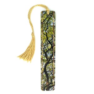 May include: A rectangular bookmark featuring a photographic print of tree branches with green leaves against a blue sky. The bookmark has a vertical border pattern of small brown and cream squares and includes a gold-colored twisted cord tassel.