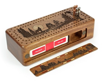 May include: A wooden cribbage board with a wolf and forest scene engraved on the surface. The board has a slot for cards and comes with a set of pegs. The board is made of dark wood and has a natural finish.