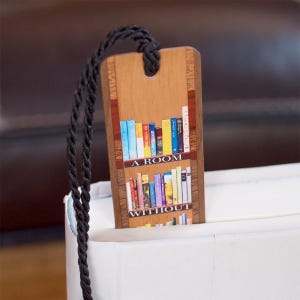 May include: A rectangular wooden bookmark featuring a printed illustration of two shelves filled with colorful books. The text A ROOM WITHOUT is printed across the shelves. A black braided cord is attached through a hole at the top of the bookmark.