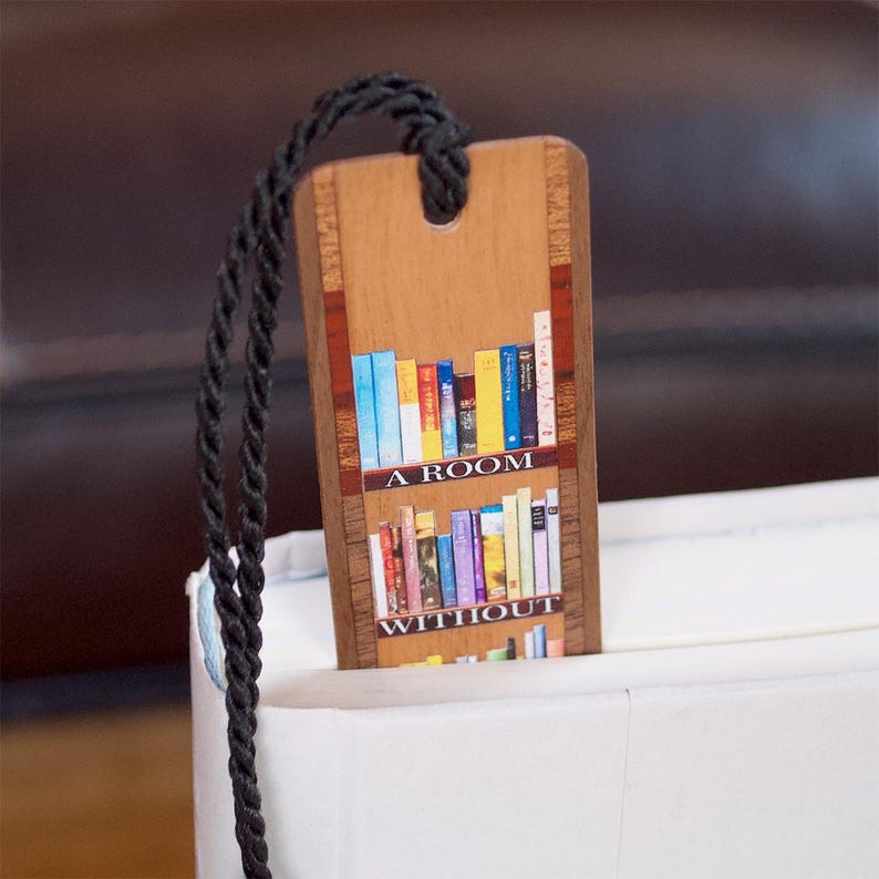 May include: A rectangular wooden bookmark featuring a printed illustration of two shelves filled with colorful books. The text A ROOM WITHOUT is printed across the shelves. A black braided cord is attached through a hole at the top of the bookmark.